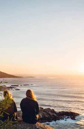 Rear view of female friends sitting at beach against clear sky during sunset
