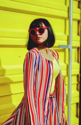 Woman in bright colors and red sunglasses posing against a yellow wall