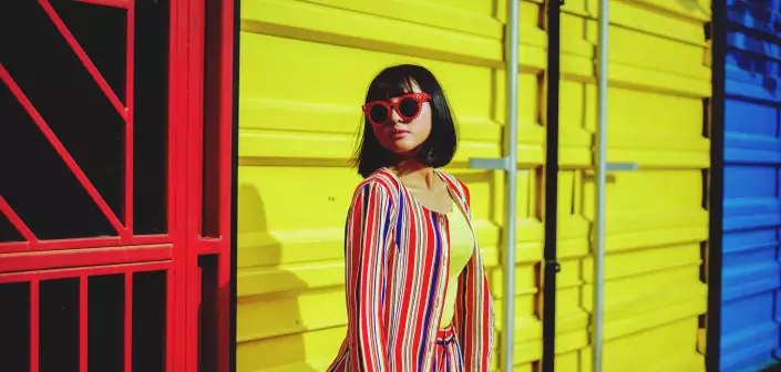 Woman in bright colors and red sunglasses posing against a yellow wall