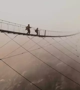 Wide shot of two people walking across suspended bridge high in the clouds